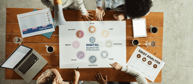 Overleg aan een houten tafel met een poster over Digital Marketing Strategies, laptop en SWOT-analyse.