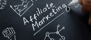 Hand schrijft "Affiliate Marketing" met een witte pen op een zwart oppervlak met schetsen van grafieken en netwerken.