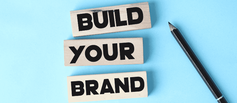 Houten blokken met de tekst "Build Your Brand" op een blauwe achtergrond, naast een potlood.