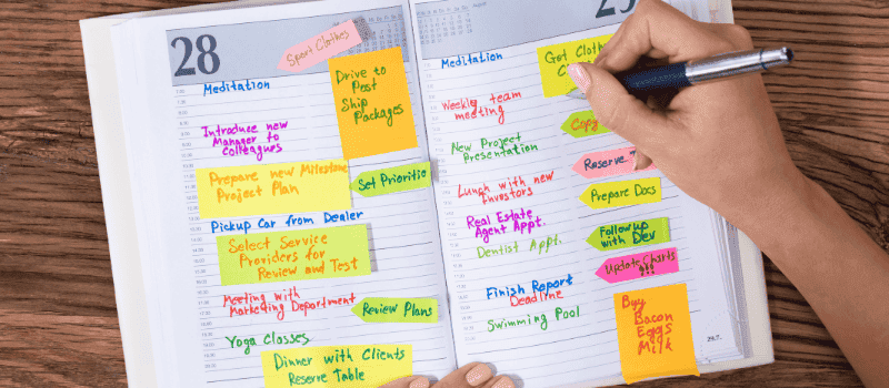 Open dagplanner met kleurrijke plaknotities en een hand die met een pen schrijft op een houten tafel.
