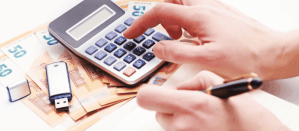 Hand gebruikt een rekenmachine op eurobiljetten, met een USB-stick en een pen op de achtergrond.