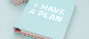 Lichtblauw notitieboek met de tekst "I Have a Plan" en een roze binnenkant op een pastelblauwe achtergrond.