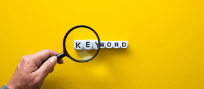 Hand houdt een vergrootglas over witte blokken met het woord "Keyword" op een gele achtergrond.