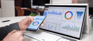 Hand houdt smartphone met marketingdashboard terwijl gegevens op een laptop worden weergegeven.