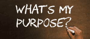 Hand schrijft met krijt de tekst "What's My Purpose?" op een donker krijtbord, focus op zelfreflectie.