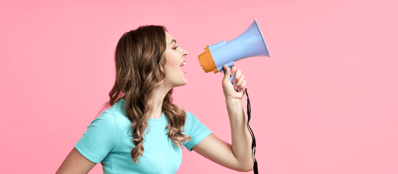 Jonge vrouw met lang bruin haar roept in een blauwe megafon tegen een roze achtergrond.
