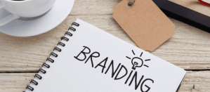 Handgeschreven woord "BRANDING" met een gloeilampillustratie in notitieboek naast een kop koffie.